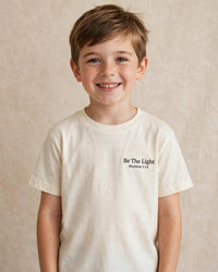 Thumbnail for Child wearing the cream Kids Be The Light Tee - Be The Light, Matthew 5:14 print over the heart